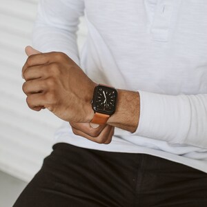 May include: A black smartwatch with a rectangular face and a tan leather strap. The watch display features a dark background with white hour and minute hands, showing a classic analog clock interface with a small red accent near the bottom.