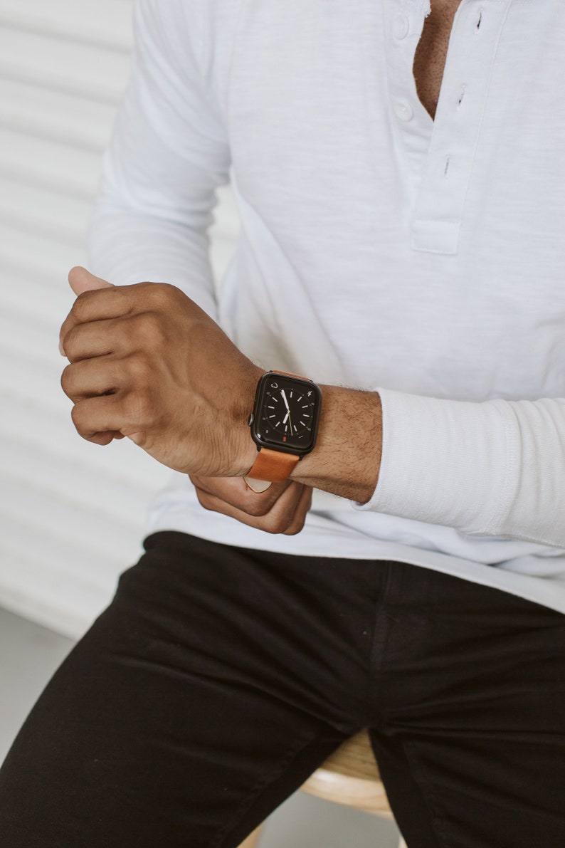 May include: A black smartwatch with a rectangular face and a tan leather strap. The watch display features a dark background with white hour and minute hands, showing a classic analog clock interface with a small red accent near the bottom.