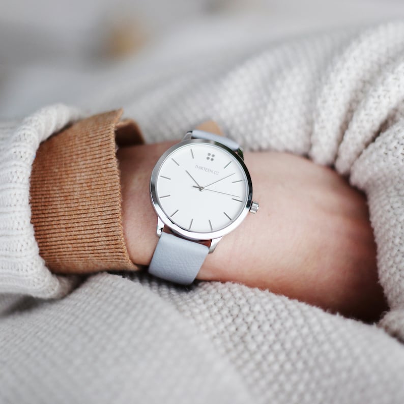 May include: A silver watch with a white face and a light blue leather band. The watch face has the text "THIRTEEN" and a small logo.
