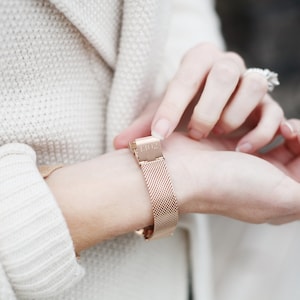 May include: A rose gold metal watch band with a mesh design. The band has a clasp with the number "1302" engraved on it.
