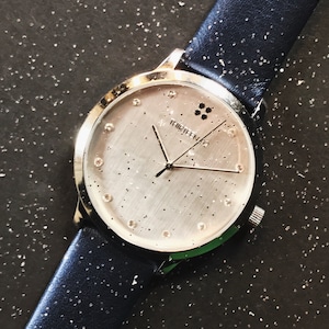 Puede incluir: Un reloj de pulsera plateado con una correa de cuero azul marino. La esfera del reloj es plateada con peque&ntilde;os puntos dispersos e indicadores de hora de cristal. El reloj tiene la palabra "thirteen" impresa en la esfera.