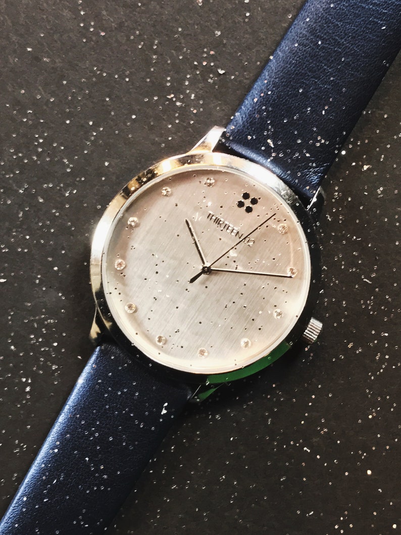 Puede incluir: Un reloj de pulsera plateado con una correa de cuero azul marino. La esfera del reloj es plateada con peque&ntilde;os puntos dispersos e indicadores de hora de cristal. El reloj tiene la palabra "thirteen" impresa en la esfera.