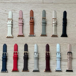 May include: A collection of 14 different colored leather watch bands for Apple Watch. The bands are arranged in two rows of seven, with each band featuring a different color, including white, pink, brown, gray, gold, black, red, dark brown, light gray, burgundy, white, and gray.