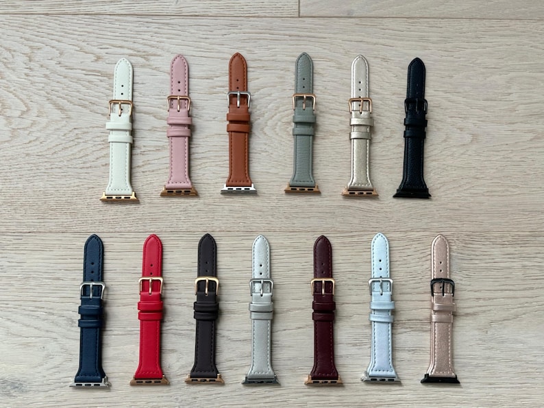 May include: A collection of 14 different colored leather watch bands for Apple Watch. The bands are arranged in two rows of seven, with each band featuring a different color, including white, pink, brown, gray, gold, black, red, dark brown, light gray, burgundy, white, and gray.