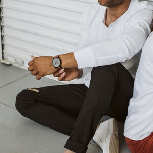 Pode incluir: Uma pessoa vestindo uma camisa branca de manga comprida, jeans preto e t&ecirc;nis branco. A pessoa est&aacute; sentada em um ch&atilde;o cinza e tem um rel&oacute;gio no pulso esquerdo.