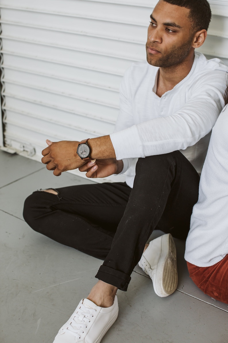 Pode incluir: Uma pessoa vestindo uma camisa branca de manga comprida, jeans preto e t&ecirc;nis branco. A pessoa est&aacute; sentada em um ch&atilde;o cinza e tem um rel&oacute;gio no pulso esquerdo.