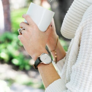 Puede incluir: Un reloj de pulsera plateado con una correa de cuero negro. El reloj tiene una esfera redonda con un dise&ntilde;o minimalista. La persona lleva un su&eacute;ter blanco y sostiene una taza blanca.