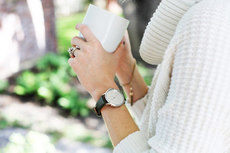 Puede incluir: Un reloj de pulsera plateado con una correa de cuero negro. El reloj tiene una esfera redonda con un dise&ntilde;o minimalista. La persona lleva un su&eacute;ter blanco y sostiene una taza blanca.