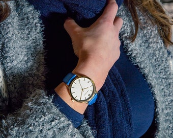 May include: A gold watch with a white face and a blue leather strap. The watch is on a person's wrist.