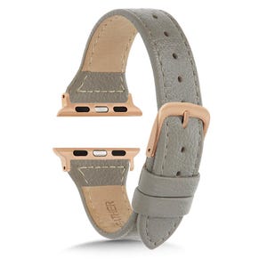 May include: A grey leather watch strap with rose gold connectors and a buckle. The strap is made of textured leather and has a light beige interior. The rose gold connectors are for a smartwatch.