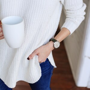 Puede incluir: Una persona con un su&eacute;ter de punto blanco texturizado con cuello alto y vaqueros azul oscuro. Una taza blanca se sostiene en una mano y un reloj con esfera plateada y correa de cuero negro en la mu&ntilde;eca.