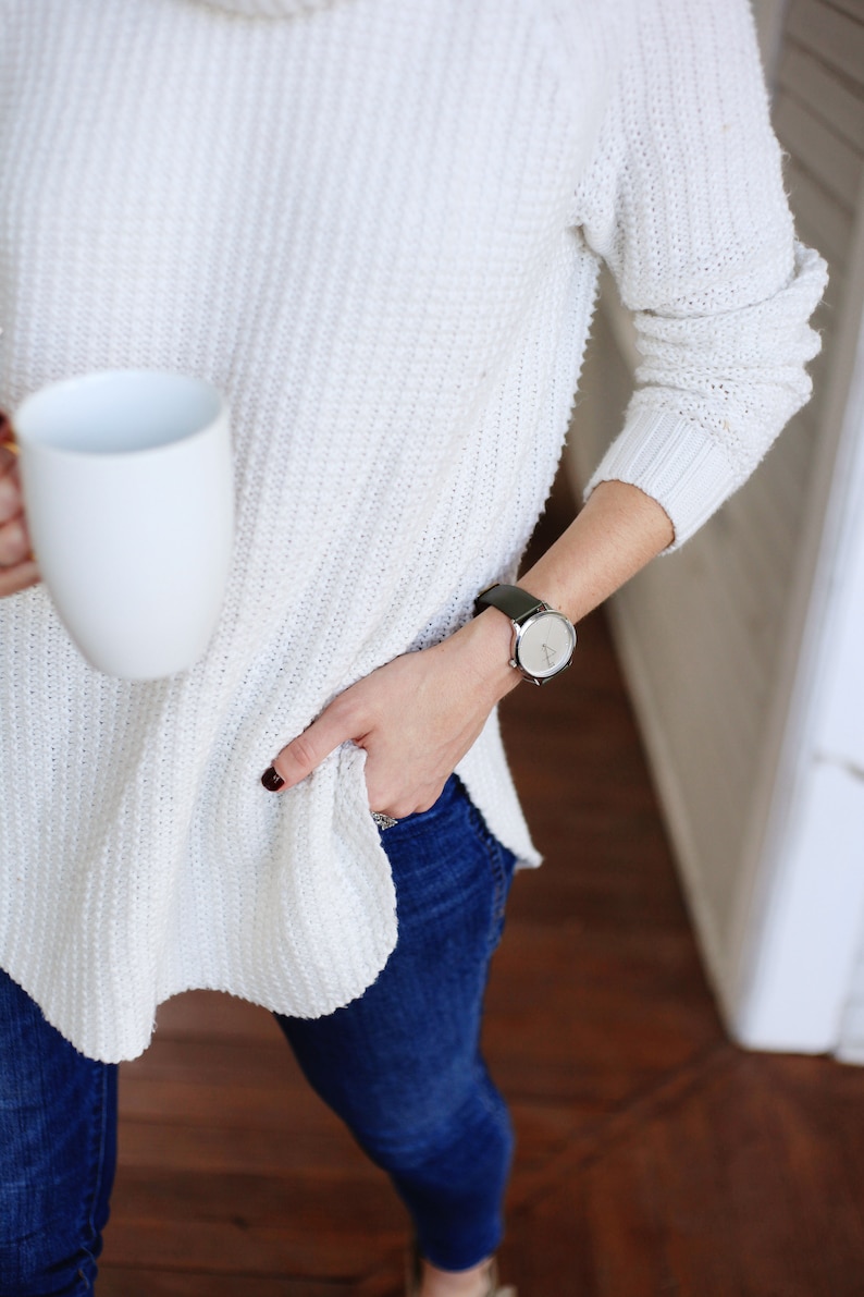Puede incluir: Una persona con un su&eacute;ter de punto blanco texturizado con cuello alto y vaqueros azul oscuro. Una taza blanca se sostiene en una mano y un reloj con esfera plateada y correa de cuero negro en la mu&ntilde;eca.