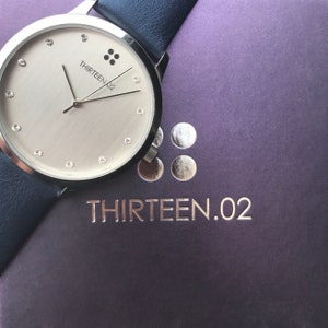 Puede incluir: Un reloj plateado con esfera clara, con marcadores tipo diamante y el texto "THIRTEEN.02". El reloj tiene una correa de cuero azul oscuro. Descansa sobre una superficie p&uacute;rpura con el nombre de la marca en relieve.