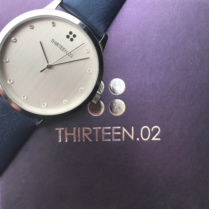 Puede incluir: Un reloj plateado con esfera clara, con marcadores tipo diamante y el texto "THIRTEEN.02". El reloj tiene una correa de cuero azul oscuro. Descansa sobre una superficie p&uacute;rpura con el nombre de la marca en relieve.