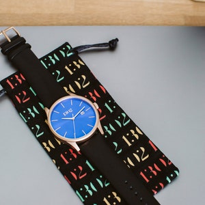 K&ouml;nnte beinhalten: Eine ros&eacute;goldene Uhr mit blauem Zifferblatt und schwarzem Lederarmband. Die Uhr befindet sich in einer schwarzen Tasche mit bunten Zahlen darauf.