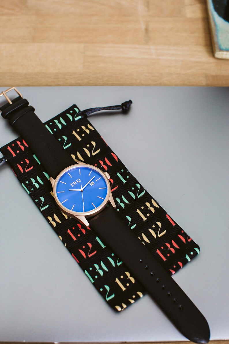 K&ouml;nnte beinhalten: Eine ros&eacute;goldene Uhr mit blauem Zifferblatt und schwarzem Lederarmband. Die Uhr befindet sich in einer schwarzen Tasche mit bunten Zahlen darauf.