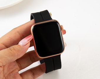May include: A rose gold smartwatch with a black screen and black silicone band. The watch has a rectangular face with rounded corners. The band is secured with a buckle. The watch is held in a hand.
