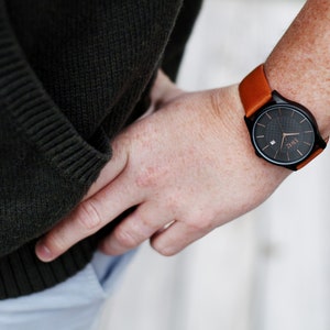 Pode incluir: Um rel&oacute;gio de pulso preto com uma pulseira de couro marrom. O mostrador do rel&oacute;gio tem um fundo preto com marcas brancas e a palavra "TIME" em letras brancas.