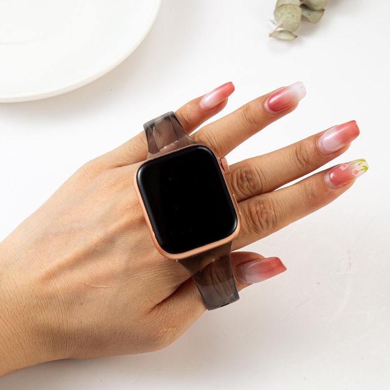 K&ouml;nnte beinhalten: Ein transparentes, durchsichtiges Armband mit einem schwarzen und grauen Muster. Das Armband ist an einer ros&eacute;goldenen Smartwatch mit einem schwarzen Bildschirm befestigt.