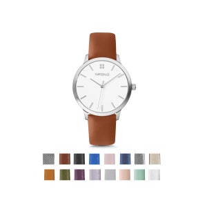 May include: A silver watch with a white face and brown leather band. The watch face has a small diamond-shaped logo with the text "THIRTEEN02" below it. The watch is shown against a white background.