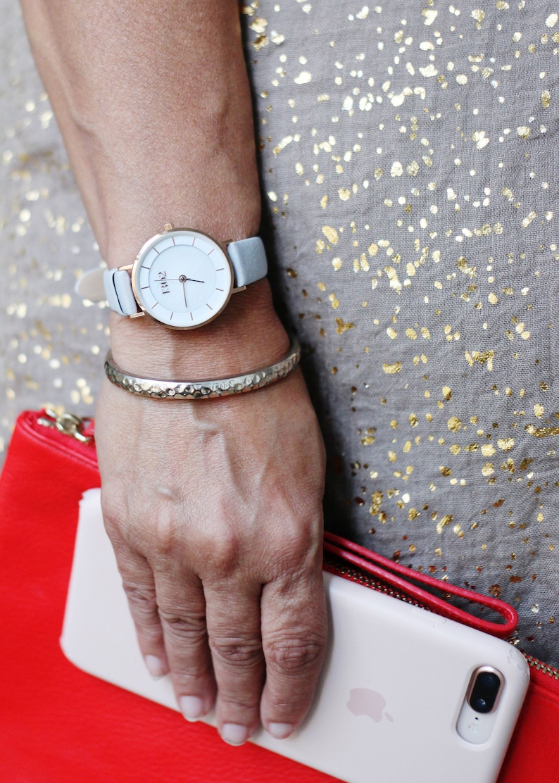 May include: A woman's wrist with a silver bangle bracelet and a white watch with a light blue leather band. The watch face has black Roman numeral markings and the word "Time" printed in black. The woman is wearing a gray dress with gold flecks.