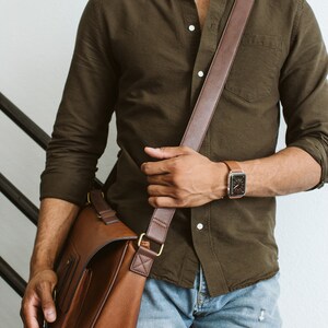 May include: An olive green button-down shirt with long sleeves, light blue denim jeans, a brown leather messenger bag with a wide shoulder strap, and a smartwatch with a brown leather band worn on the wrist.