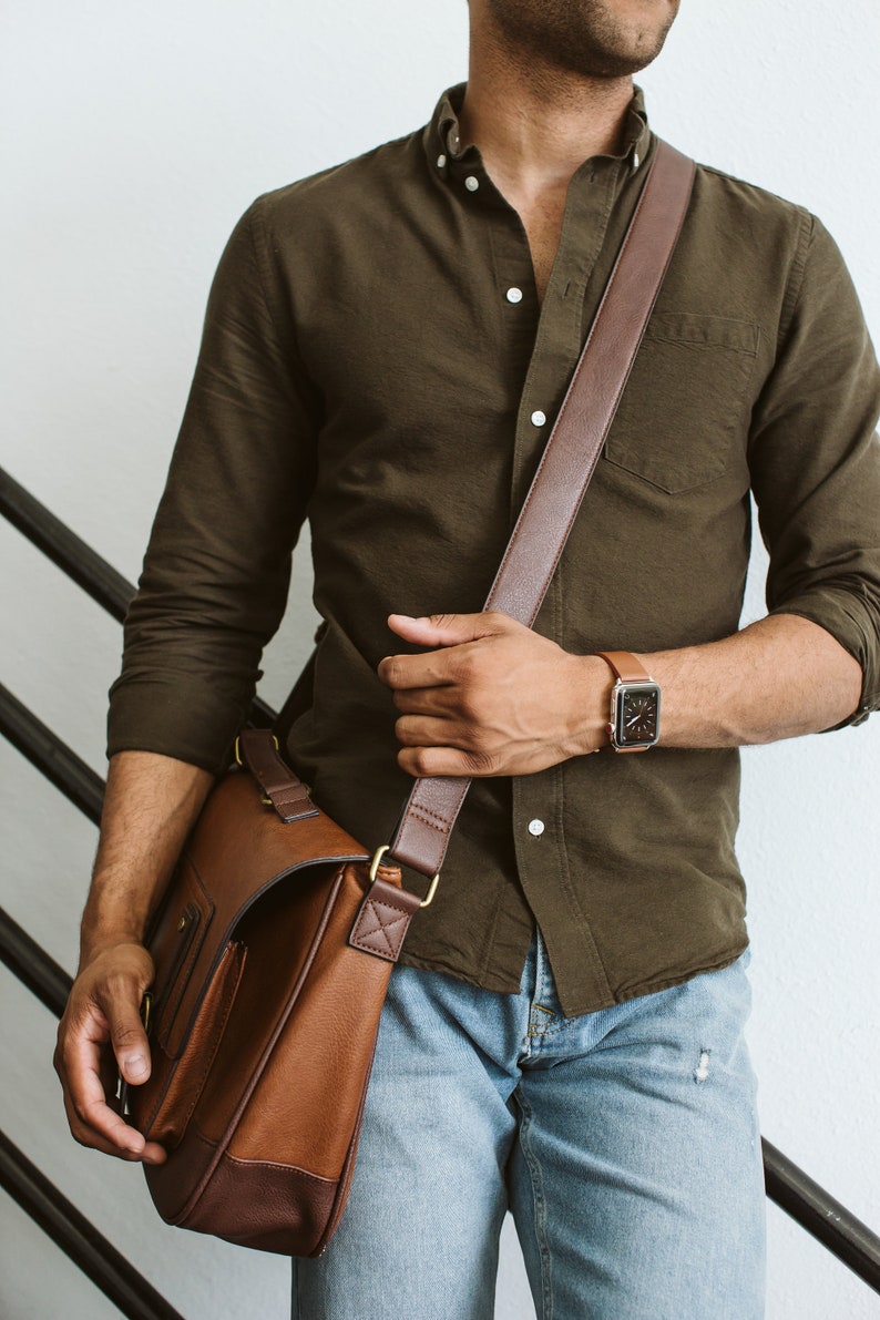 May include: An olive green button-down shirt with long sleeves, light blue denim jeans, a brown leather messenger bag with a wide shoulder strap, and a smartwatch with a brown leather band worn on the wrist.