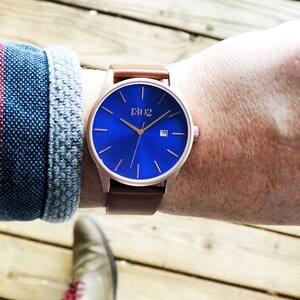 K&ouml;nnte beinhalten: Eine ros&eacute;goldene Armbanduhr mit blauem Zifferblatt und braunem Lederarmband. Das Zifferblatt hat goldene Zeiger und Markierungen, und das Datumsfenster befindet sich bei 3 Uhr. Auf dem Zifferblatt ist auch der Text "1302" aufgedruckt.