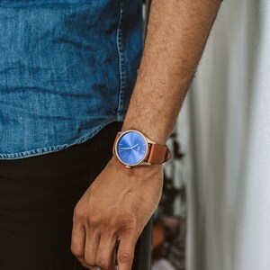 K&ouml;nnte beinhalten: Eine Person tr&auml;gt ein blaues Jeanshemd und eine schwarze Hose mit einer braunen Lederarmbanduhr mit blauem Zifferblatt.