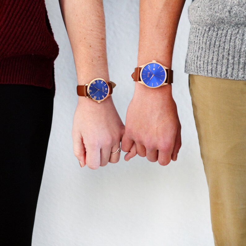 K&ouml;nnte beinhalten: Zwei Personen tragen Uhren mit braunen Lederb&auml;ndern und blauen Zifferbl&auml;ttern. Die Uhren haben goldene Akzente.