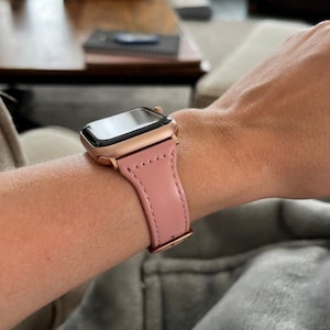 May include: A rose gold smartwatch with a pink leather strap is fastened on a wrist. The watch features a rectangular screen and a matching rose gold case. The pink strap is stitched and secured with a rose gold clasp. A fashionable wrist accessory.