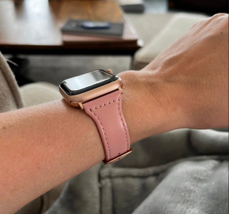 May include: A rose gold smartwatch with a pink leather strap is fastened on a wrist. The watch features a rectangular screen and a matching rose gold case. The pink strap is stitched and secured with a rose gold clasp. A fashionable wrist accessory.