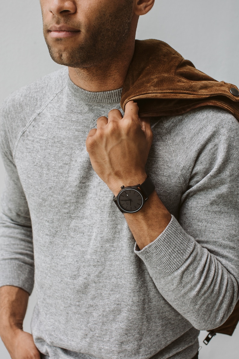 May include: A wristwatch with a black face and black band. The watch has a minimalist design and is shown on a person's wrist. The person is wearing a gray sweater and a brown suede jacket is draped over their shoulder.