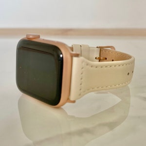May include: A rose gold smartwatch with a cream leather strap. The watch features a rectangular face with a black screen. The strap is fastened with a gold-coloured buckle. A fashionable accessory.