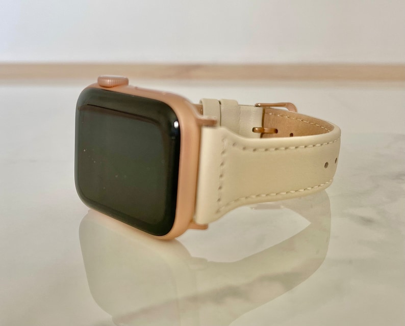 May include: A rose gold smartwatch with a cream leather strap. The watch features a rectangular face with a black screen. The strap is fastened with a gold-coloured buckle. A fashionable accessory.