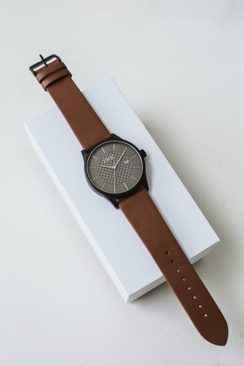 Pode incluir: Um rel&oacute;gio de pulso de couro marrom com um mostrador preto e um padr&atilde;o xadrez. O mostrador do rel&oacute;gio tem n&uacute;meros e ponteiros em ouro rosa. O rel&oacute;gio est&aacute; sobre uma superf&iacute;cie branca.