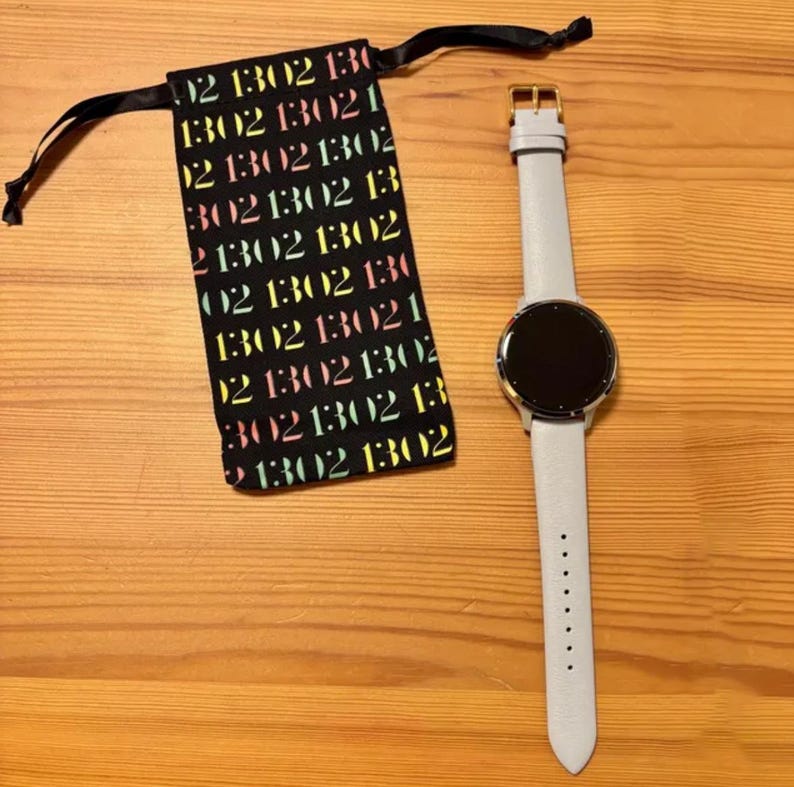 Puede incluir: Un reloj inteligente con una correa de cuero gris claro y una pantalla circular negra. Una bolsa negra con cord&oacute;n y texto colorido est&aacute; al lado del reloj. El reloj tiene una hebilla dorada.