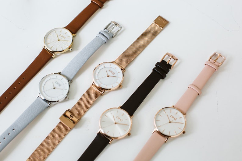 Puede incluir: Seis relojes de pulsera con esferas blancas y detalles dorados. Los relojes tienen correas de diferentes colores, incluyendo marr&oacute;n, azul claro, oro rosa, negro y rosa.