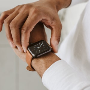 Pode incluir: Um smartwatch com uma caixa retangular cinza-escura e uma pulseira de couro castanho-claro. O mostrador digital apresenta um fundo preto com marcadores de horas brancos e ponteiros finos brancos, exibindo um design de rel&oacute;gio anal&oacute;gico.