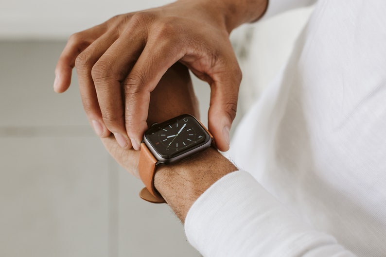 Pode incluir: Um smartwatch com uma caixa retangular cinza-escura e uma pulseira de couro castanho-claro. O mostrador digital apresenta um fundo preto com marcadores de horas brancos e ponteiros finos brancos, exibindo um design de rel&oacute;gio anal&oacute;gico.