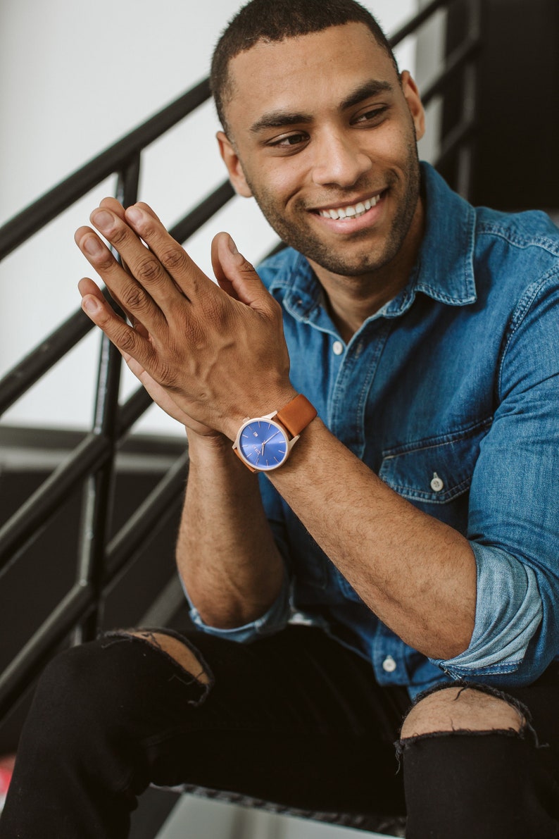 K&ouml;nnte beinhalten: Ein Mann tr&auml;gt ein blaues Jeanshemd und eine schwarze Jeans mit Destroyed-Details. Er tr&auml;gt eine Uhr mit einem braunen Lederarmband und einem blauen Zifferblatt.