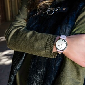 Puede incluir: Un reloj de pulsera plateado con una correa de cuero rosa claro. La esfera del reloj es plateada con manecillas y marcadores negros. El reloj se lleva en la mu&ntilde;eca, con un su&eacute;ter verde y un chaleco negro en el fondo.