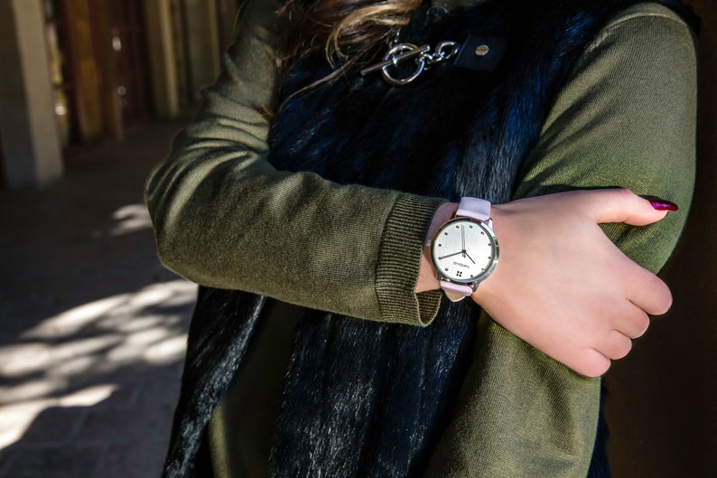 Puede incluir: Un reloj de pulsera plateado con una correa de cuero rosa claro. La esfera del reloj es plateada con manecillas y marcadores negros. El reloj se lleva en la mu&ntilde;eca, con un su&eacute;ter verde y un chaleco negro en el fondo.