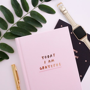 May include: A flat lay composition with a pink journal inscribed with "TODAY I AM GRATEFUL" in gold, a gold pen, a gold smartwatch with a pale strap, a black card with heart motifs, and a green leafy branch on a white background.