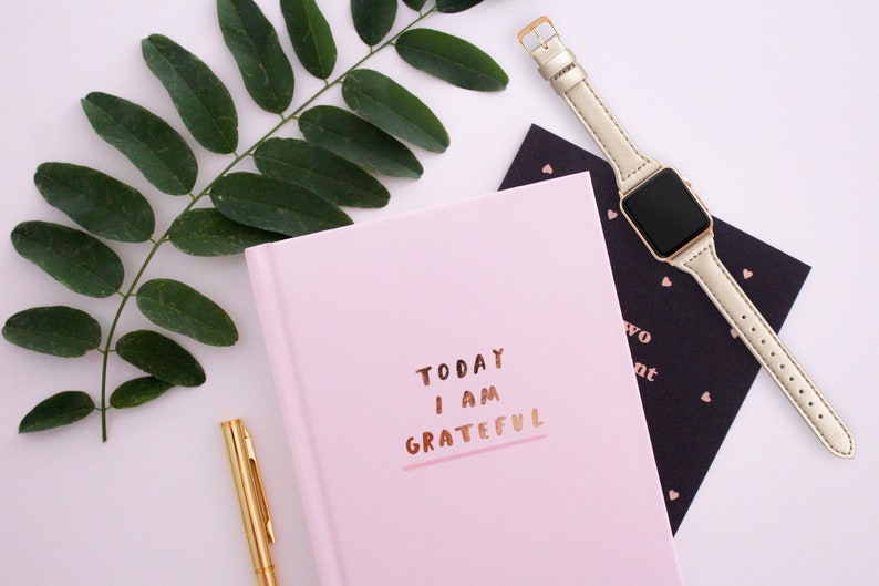 May include: A flat lay composition with a pink journal inscribed with "TODAY I AM GRATEFUL" in gold, a gold pen, a gold smartwatch with a pale strap, a black card with heart motifs, and a green leafy branch on a white background.