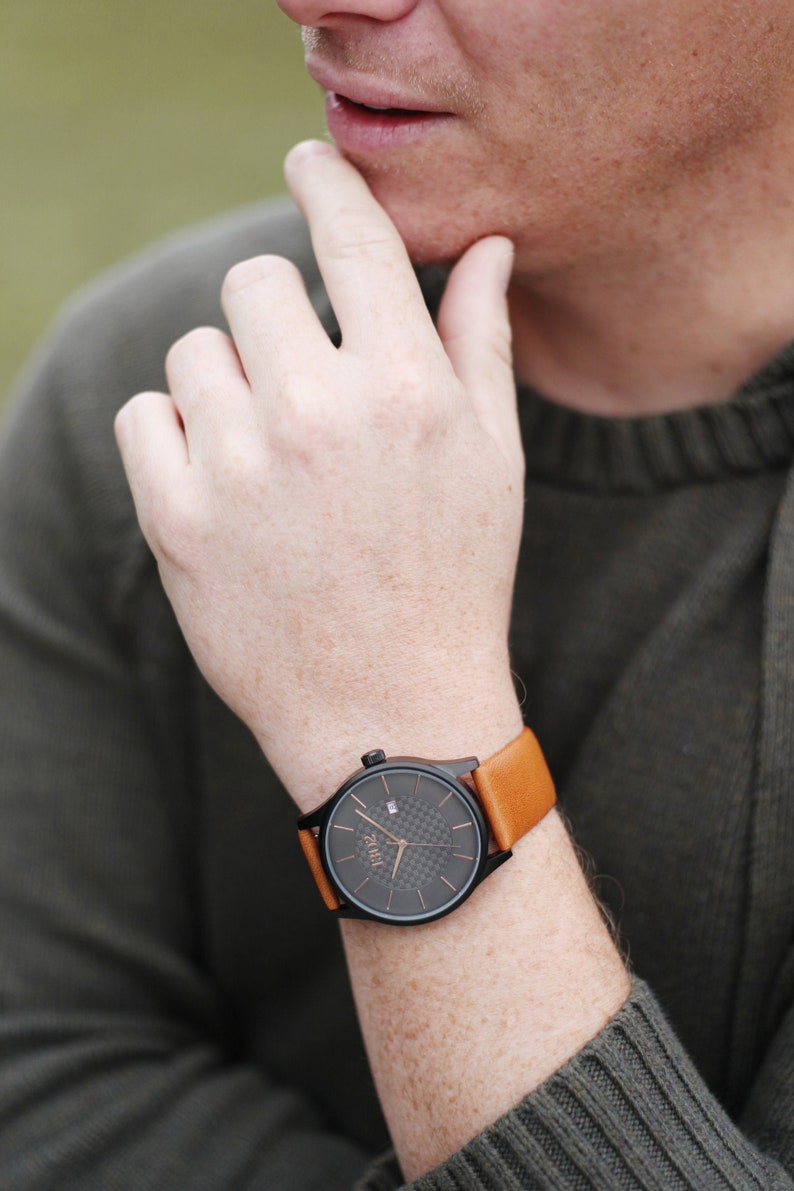 Pode incluir: Um rel&oacute;gio preto com uma pulseira de couro marrom. O mostrador do rel&oacute;gio tem um padr&atilde;o de fibra de carbono e a palavra "ZURU" impressa nele.