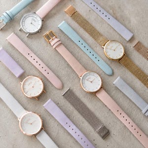 May include: A collection of analogue wristwatches with round white faces and the text "EW12" printed on the dials. The watches feature interchangeable straps in pastel pink, light blue, and lavender, alongside gold and silver-toned metal mesh bands.