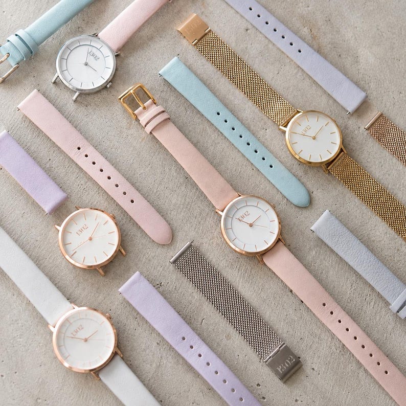 May include: A collection of analogue wristwatches with round white faces and the text "EW12" printed on the dials. The watches feature interchangeable straps in pastel pink, light blue, and lavender, alongside gold and silver-toned metal mesh bands.
