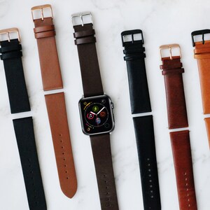 May include: A collection of leather watch straps in black, tan, and dark brown, featuring metal buckles in silver, gold, and black finishes. One dark brown strap is attached to a digital smartwatch with a circular app icon display.