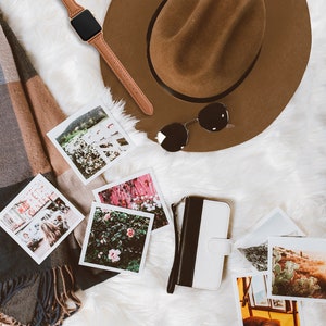 May include: A brown fedora hat, sunglasses, a brown leather watch, a black and white phone case, a scarf, and several square photographs are arranged on a white, fluffy surface. The photographs depict various scenes.